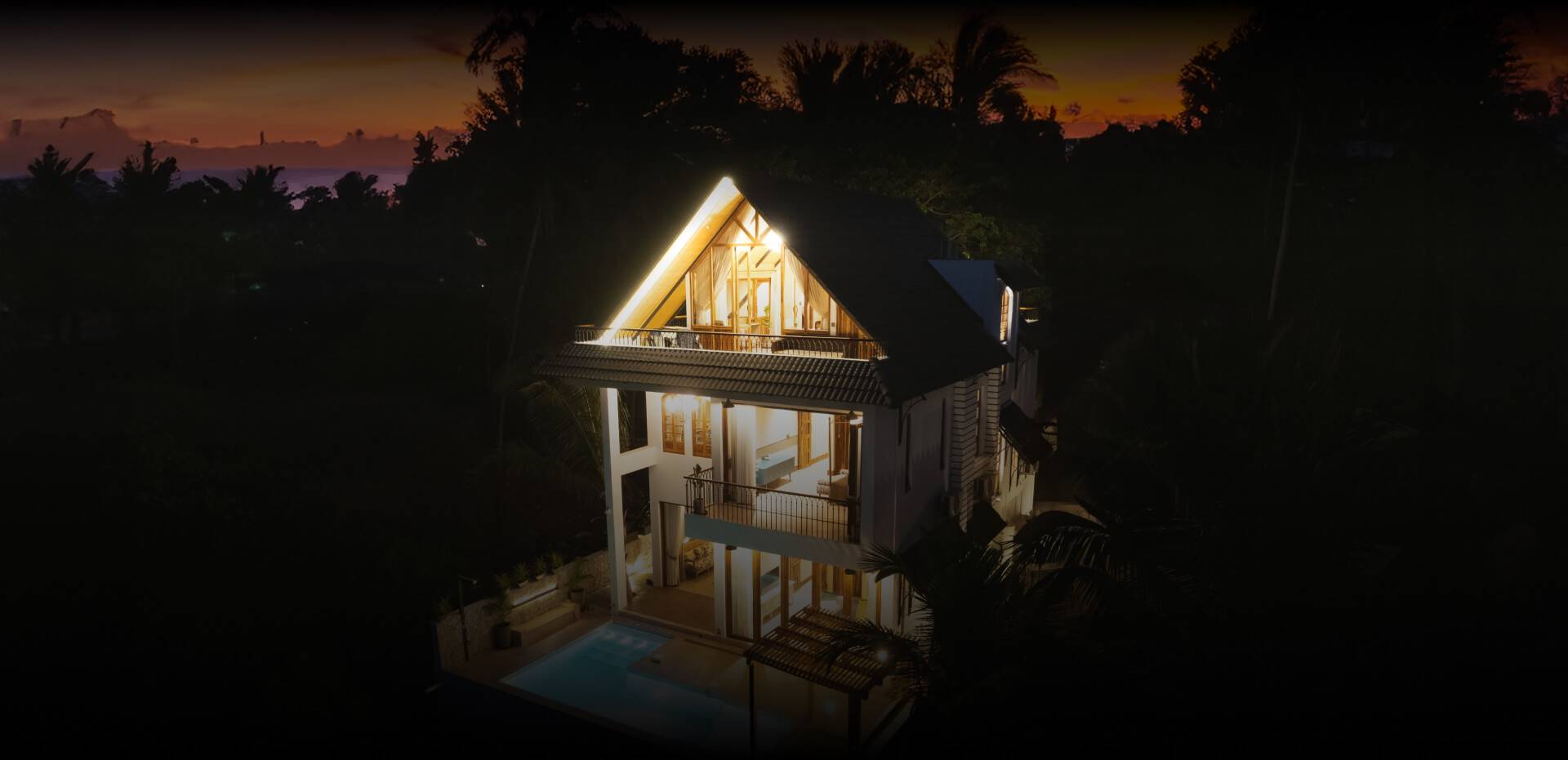 A modern three-story house illuminated at twilight, featuring large windows, balconies, and a pool, set against a backdrop of trees and a sunset sky.